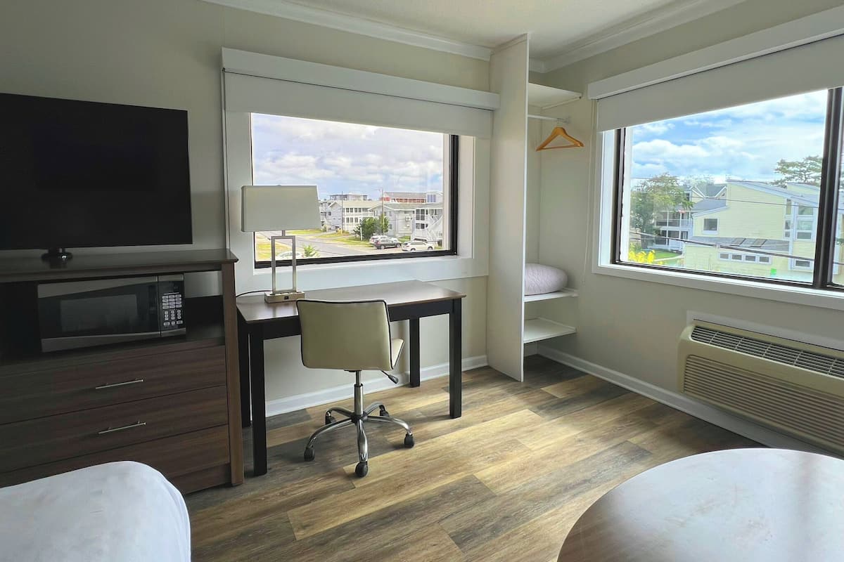 A modern hotel room with a flat-screen TV, a desk, a chair, a bed, and large windows offering a view of the street and buildings.