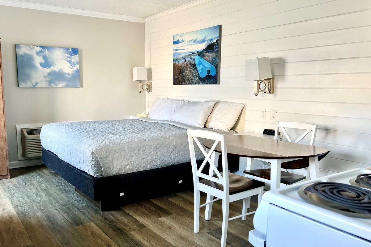 A bedroom with a bed, bedside tables, a desk with chairs, a stove, and a painting on the wall.