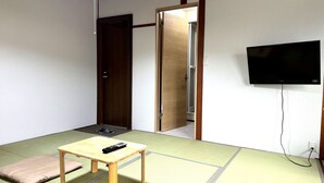 A room with tatami mats, a wooden table with a remote control, a television on the wall, and a door leading to another room. - Wafu Pension MIYAZAWA (Nozawaonsen)