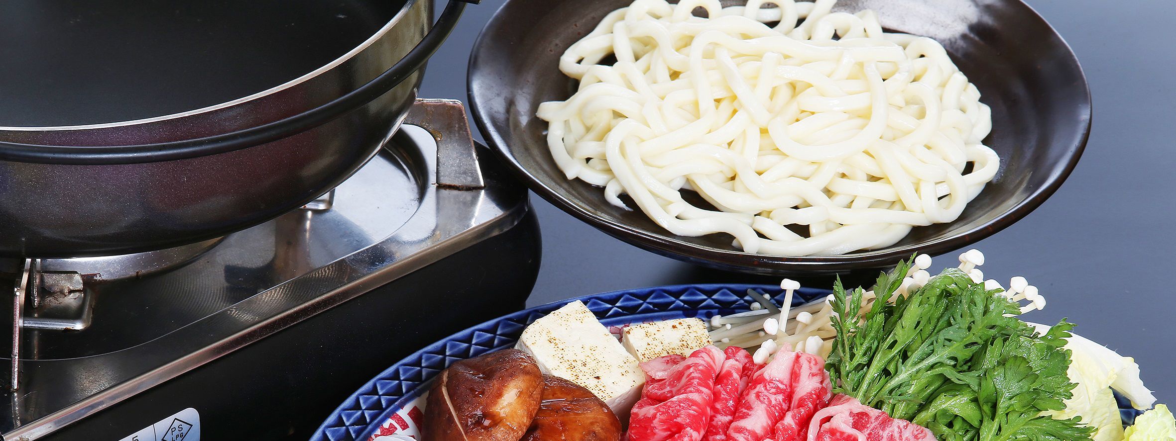 Un ensemble pour préparer une fondue, comprenant un récipient, des nouilles et divers ingrédients comme de la viande et des légumes.