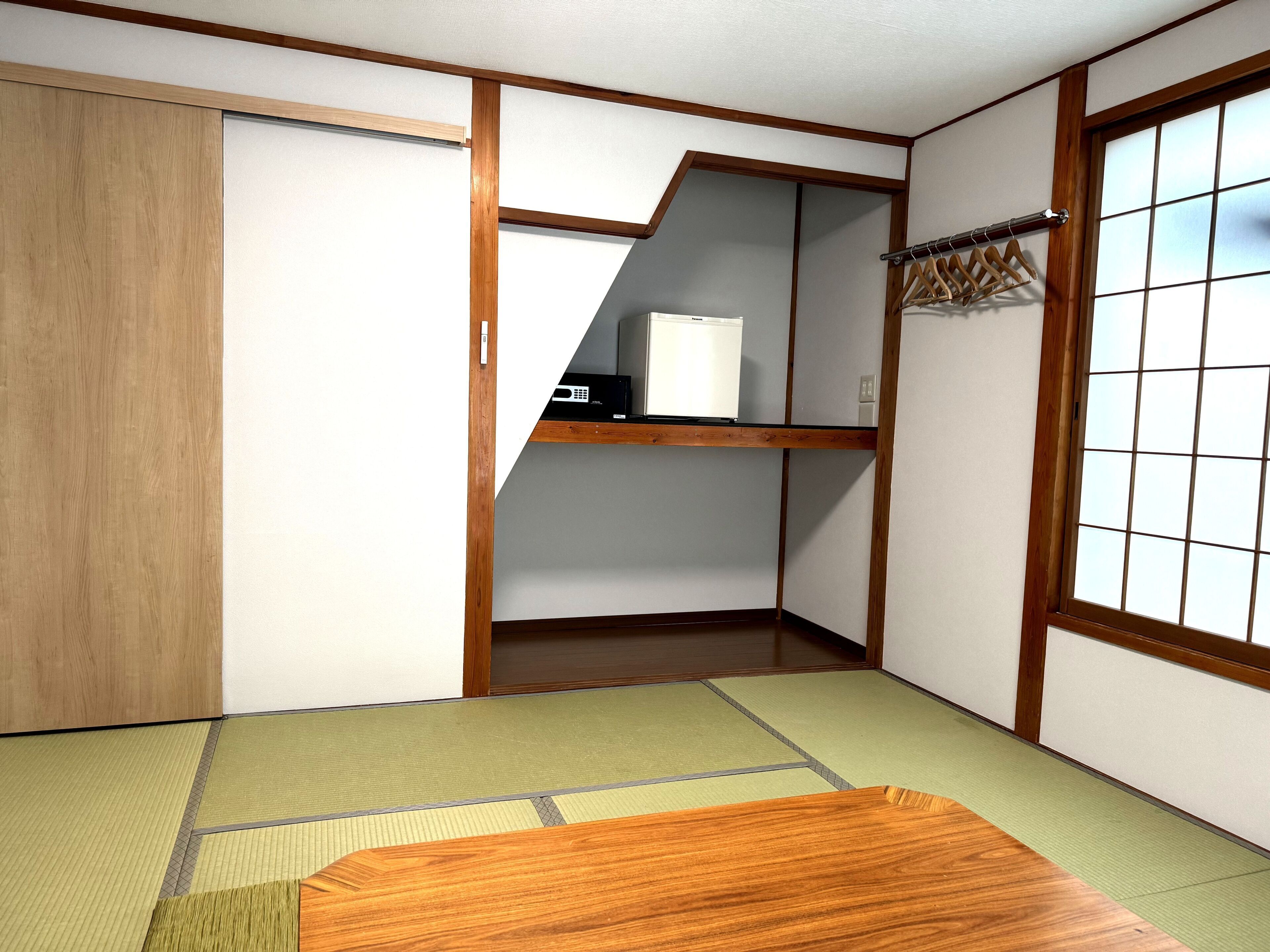 Una habitación japonesa tradicional con suelo de tatami, puertas correderas, un estante con un electrodoméstico blanco y una ventana con diseño de cuadrícula.