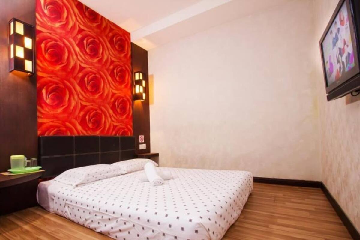 A hotel room with a bed, a wall with a red rose pattern, a wooden floor, and a wall-mounted TV.