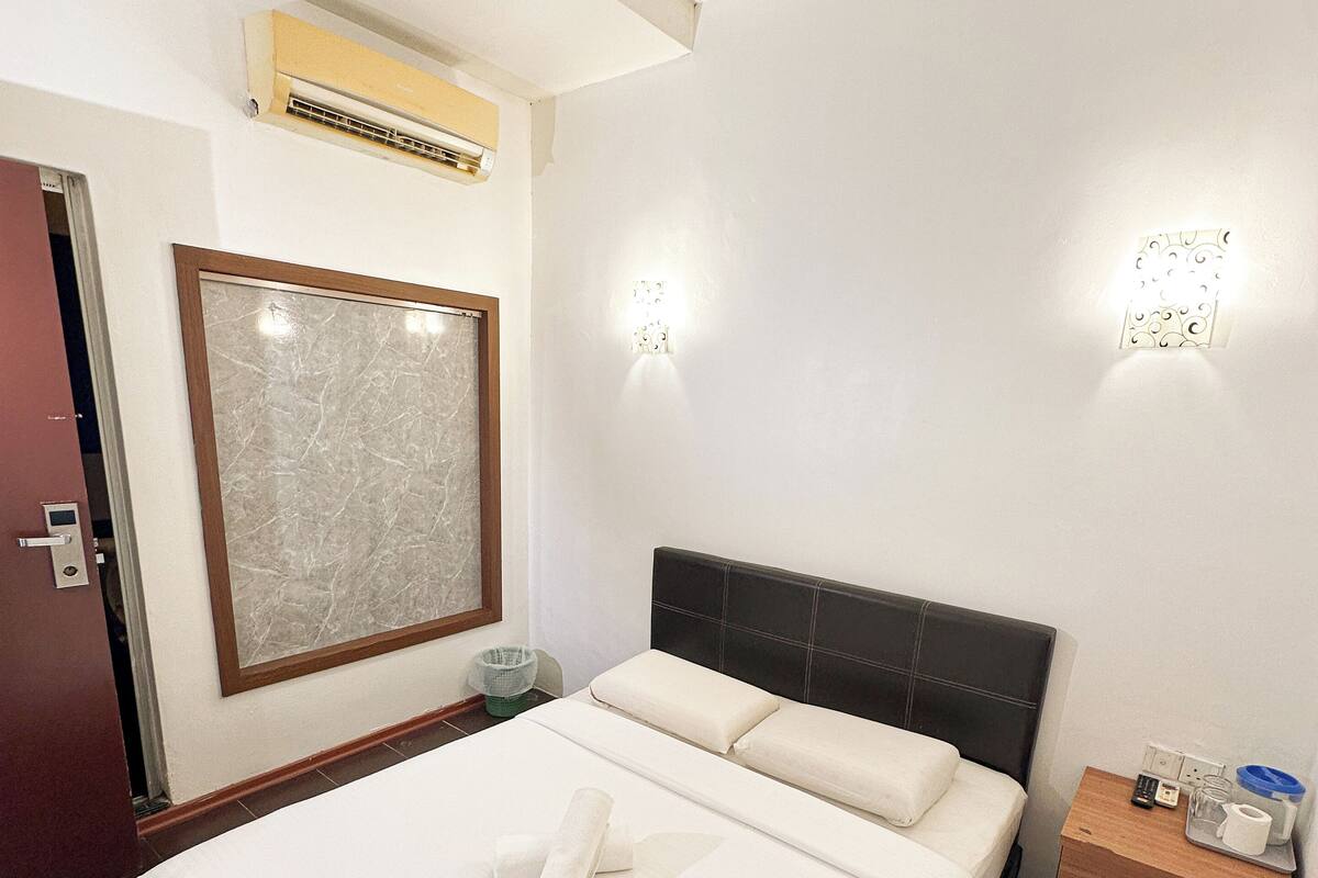 A hotel room with a large bed, a headboard, a bedside table with a phone and a cup, and an air conditioning unit on the wall.