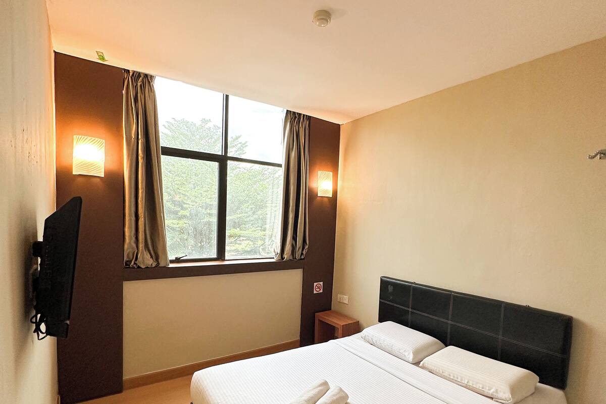 A hotel room with a bed, a TV mounted on the wall, a window with curtains, and a night light.