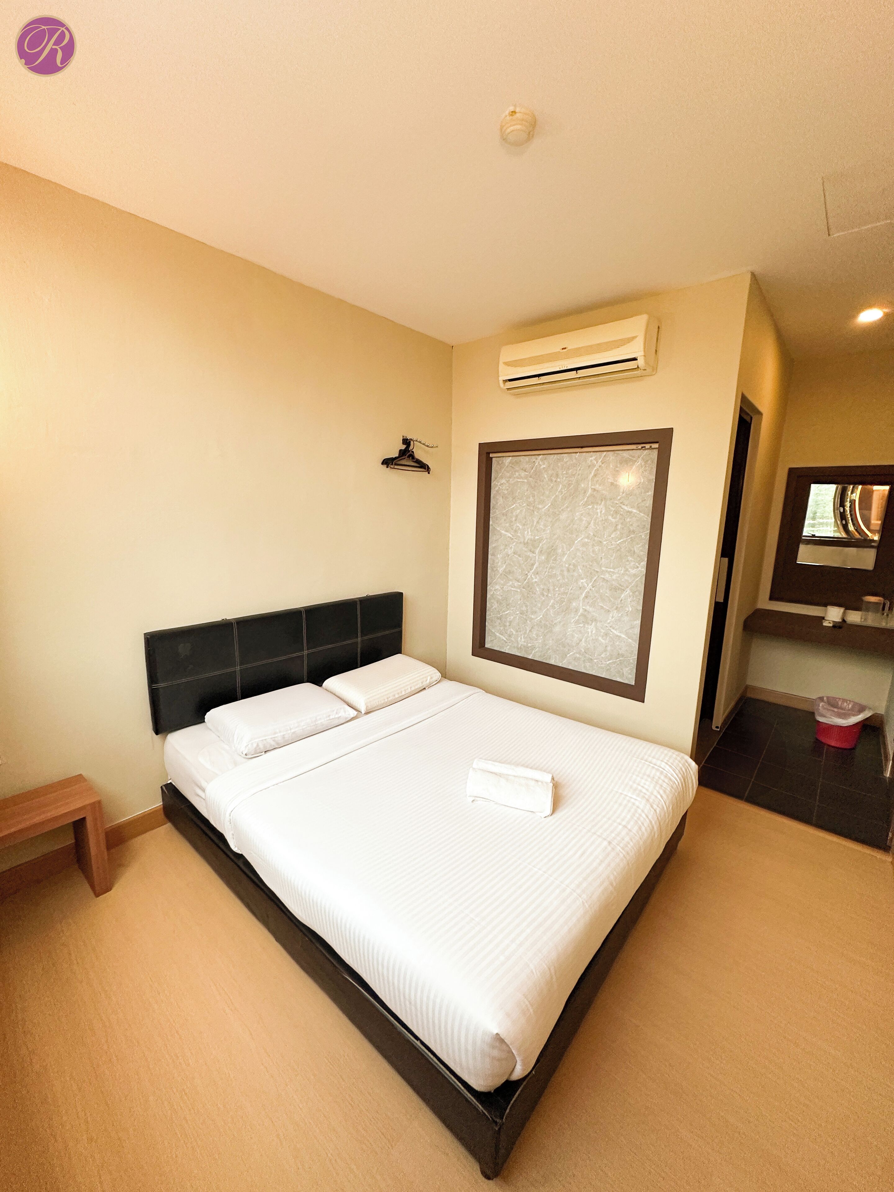 A neatly made bed with white linens, a small wooden nightstand, a wall-mounted air conditioner, and a bathroom with a mirror and sink.
