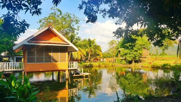 A wooden house with a balcony, situated by a calm pond, surrounded by lush greenery and trees. - Wanlapa Farmstay (Lop Buri)