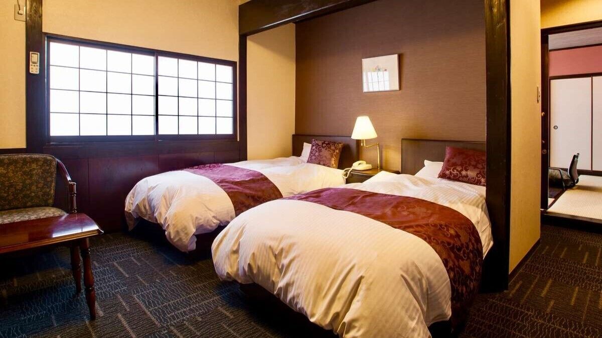 A hotel room with two beds, a chair, a bedside table with a lamp, and a window.