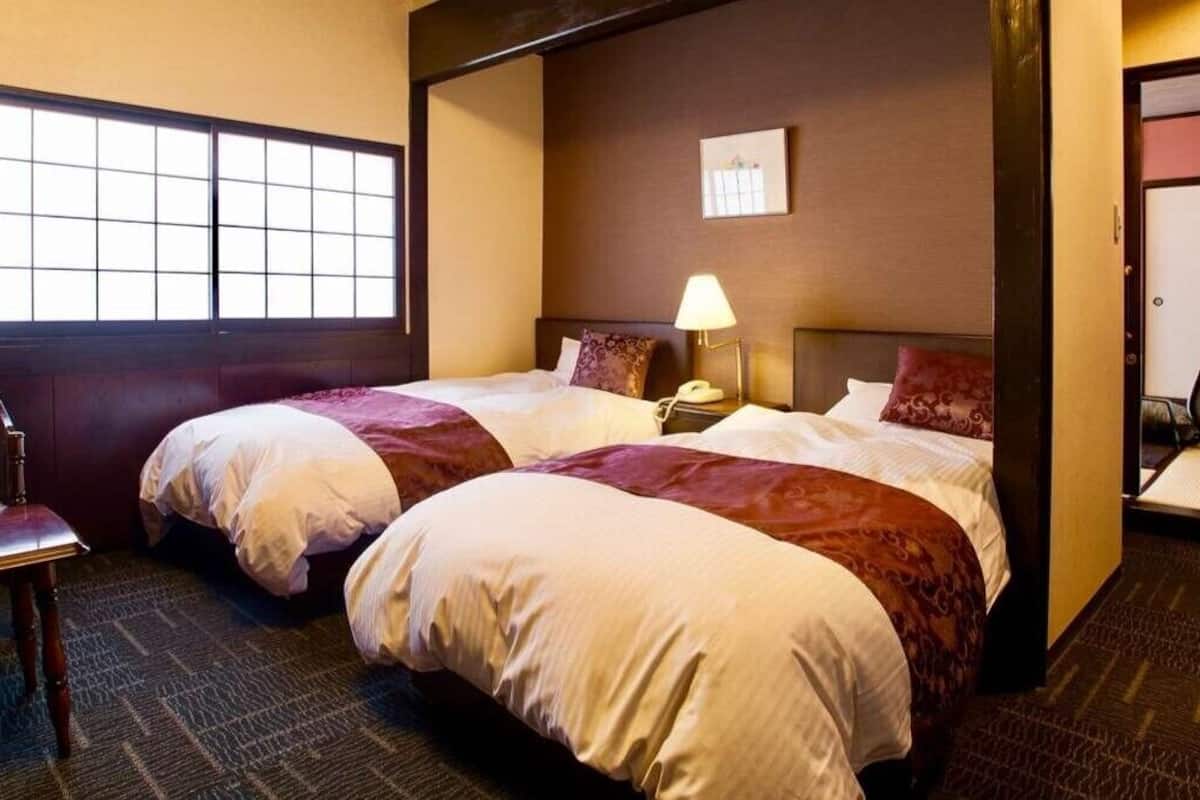 Quarto de hotel com duas camas, uma cadeira, mesa de cabeceira com abajur e uma janela.