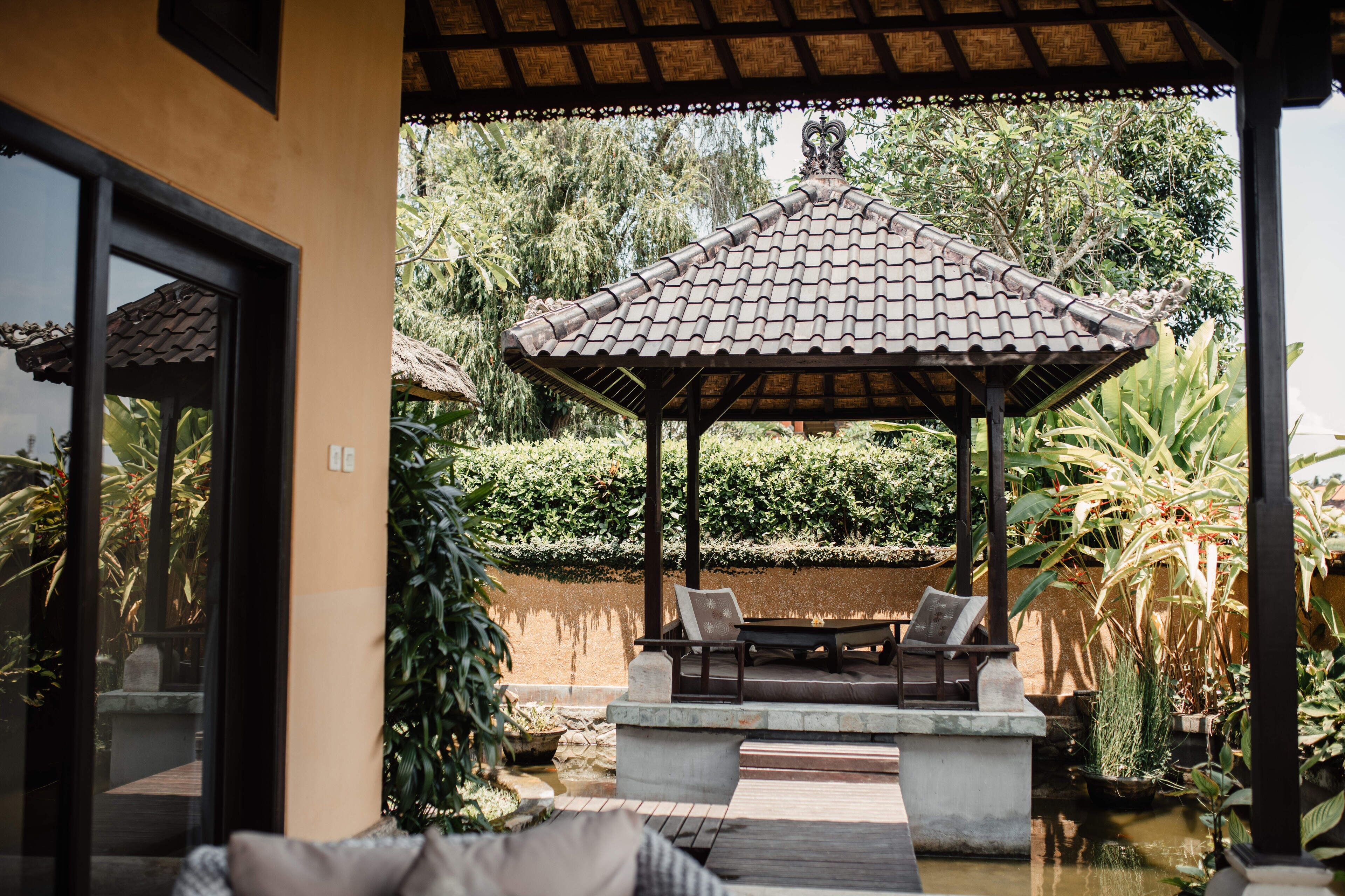A covered outdoor seating area with a tiled roof, surrounded by lush greenery and a small water feature.