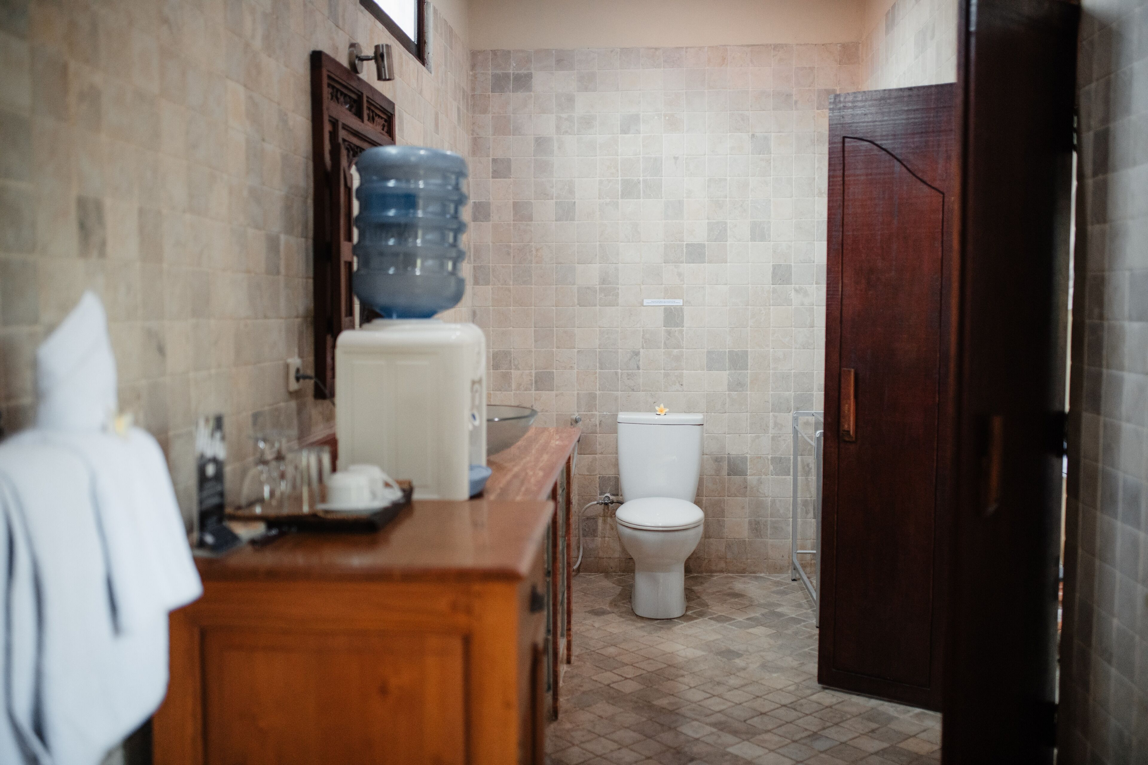 Un baño con tocador de madera, un dispensador grande de agua, un inodoro y una puerta de madera oscura.