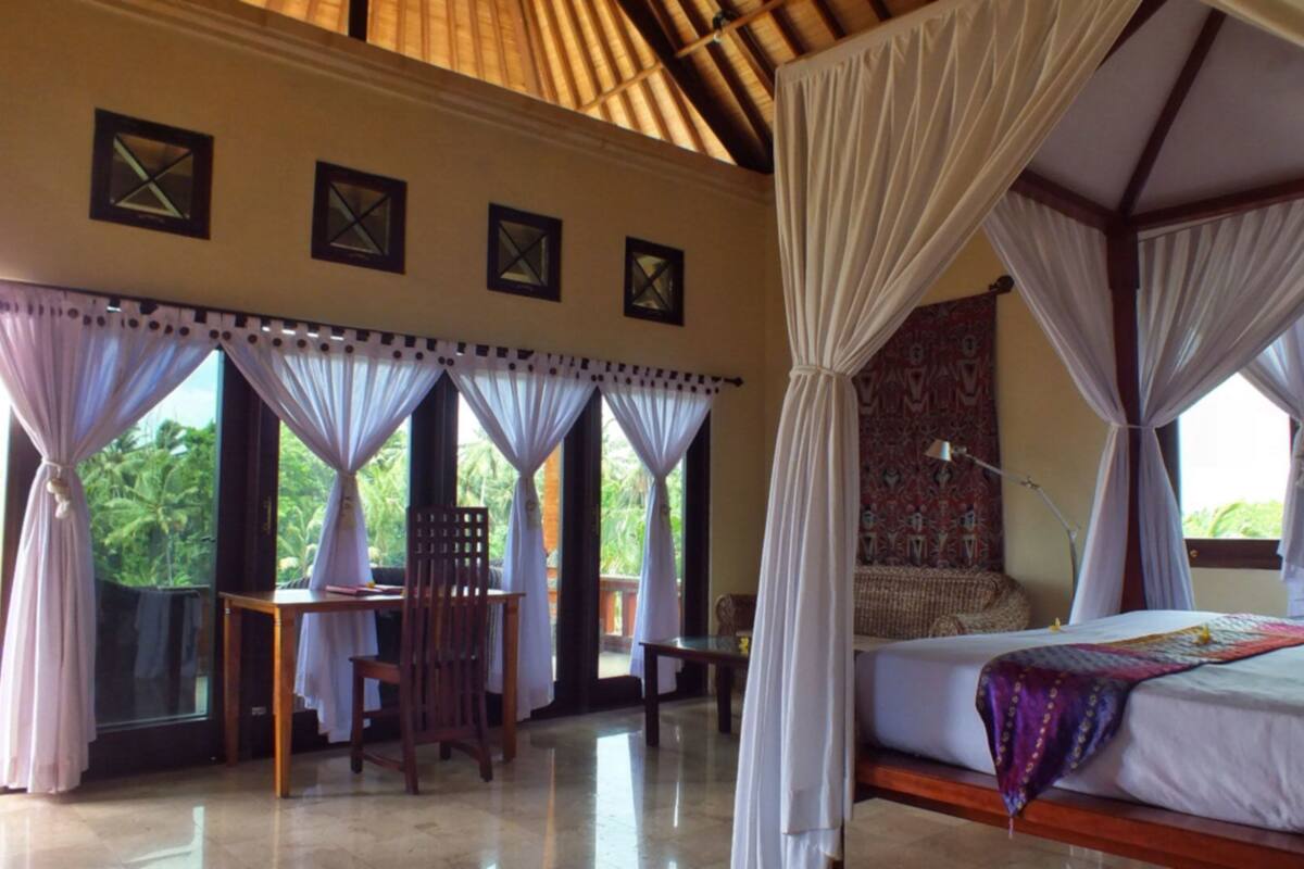 A room with a bed, a table, and a chair, with windows and curtains.