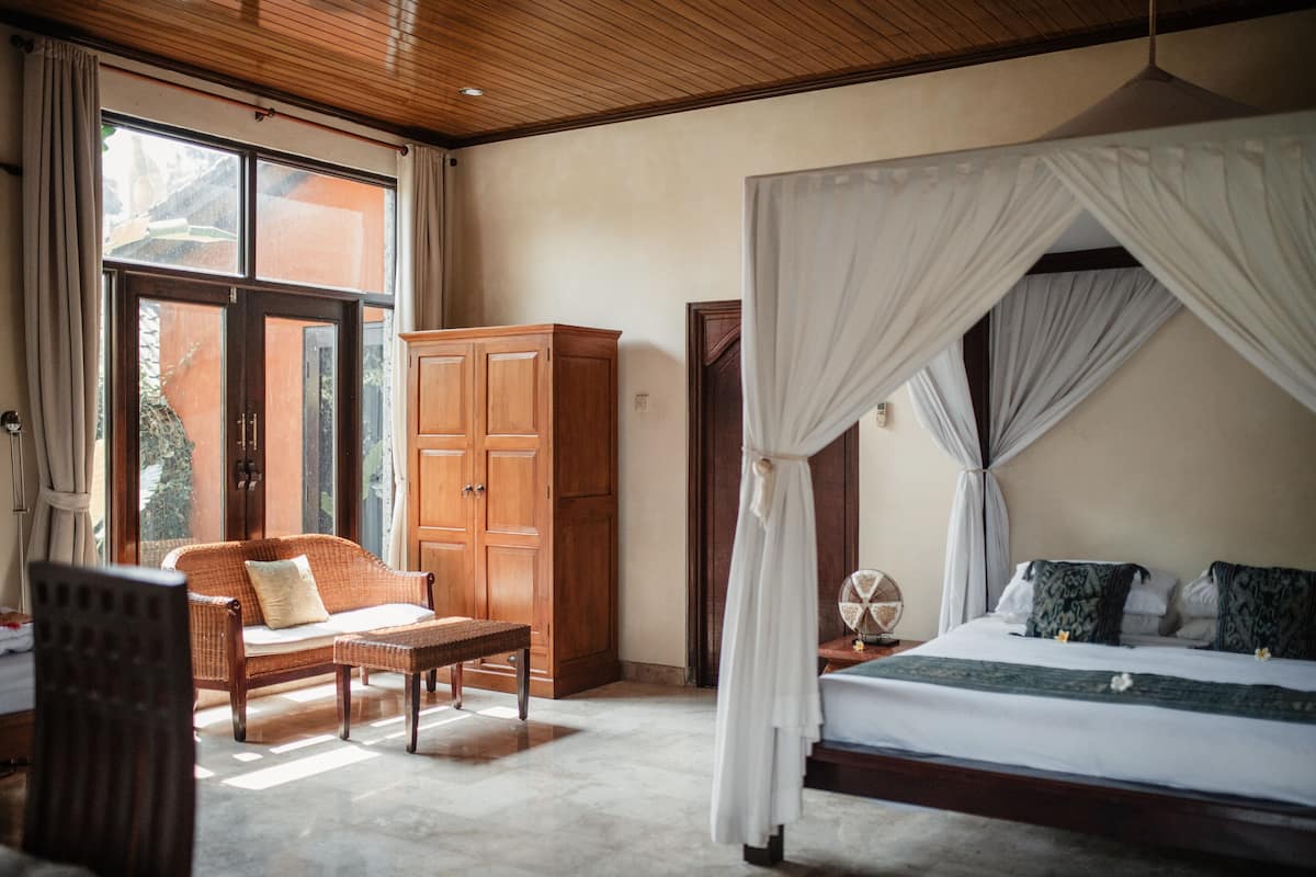 A bedroom with a four-poster bed, wooden furniture, and a view through a large window.