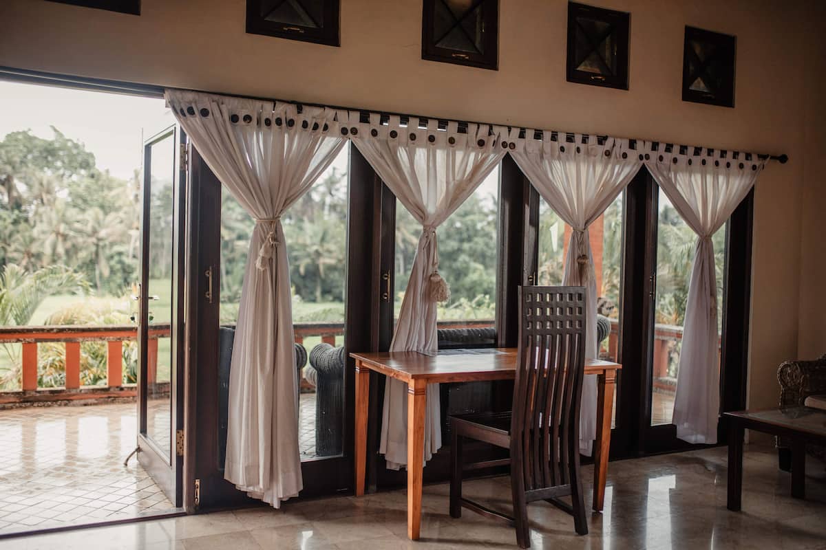 A room with a wooden table and chairs, a balcony with a railing, and a view of greenery outside.