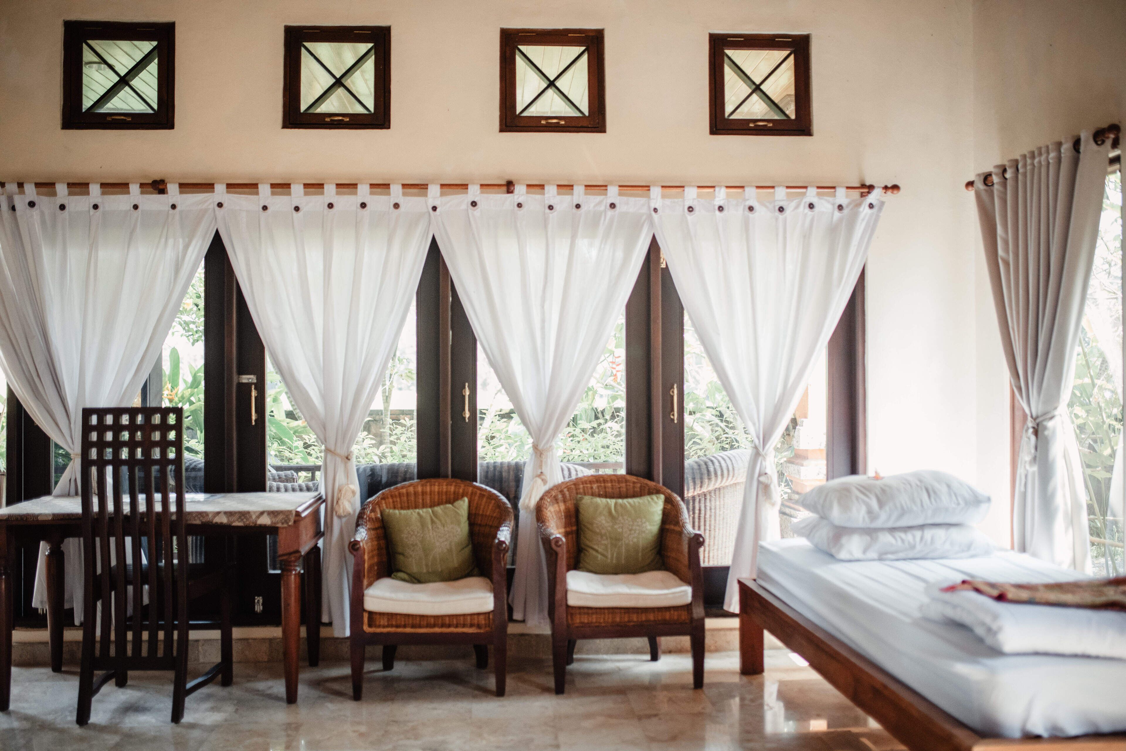 A room with a bed, two wicker chairs, a table, and a window with white curtains.
