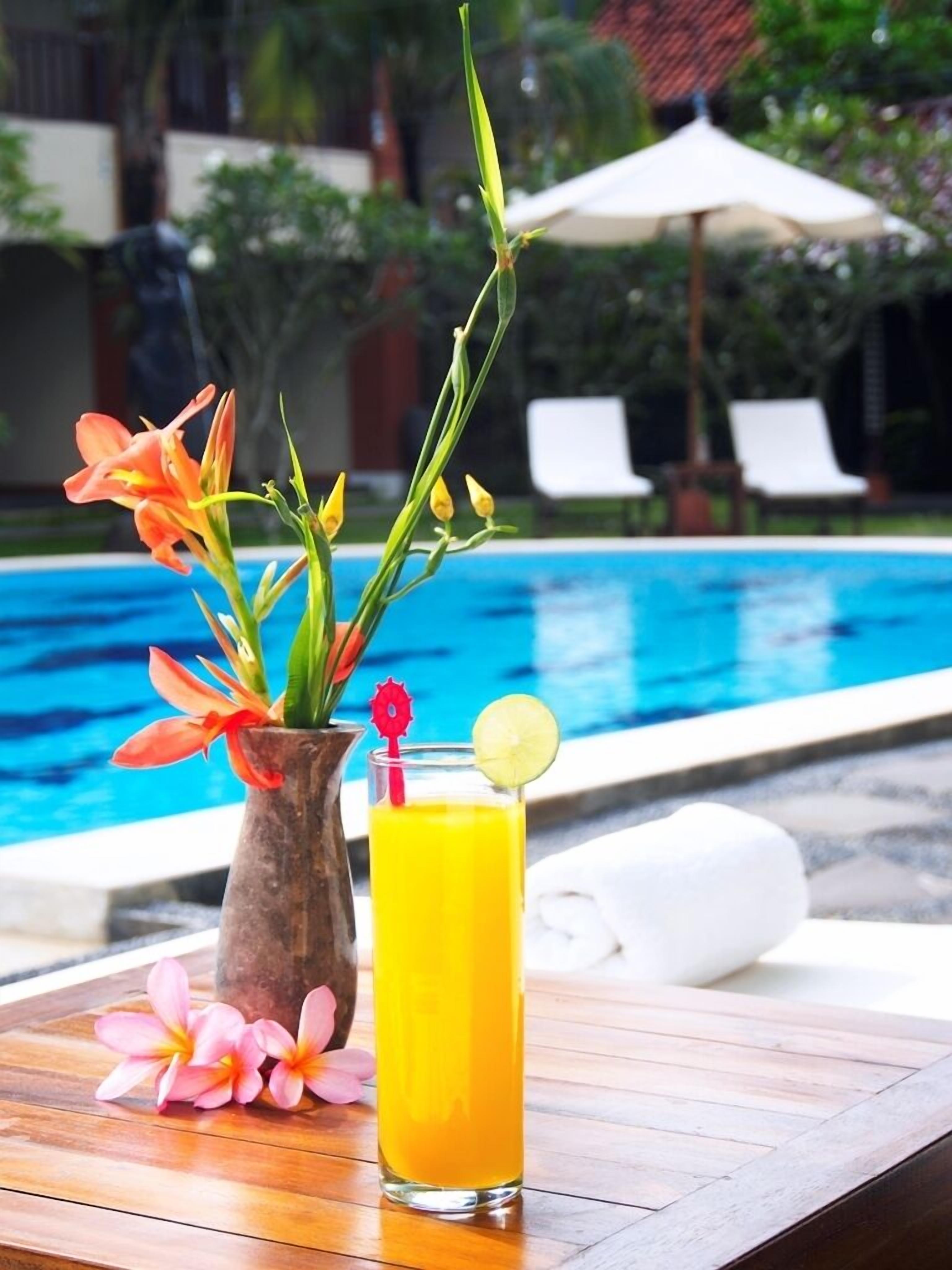 Escena junto a la piscina con una mesa de madera, un jarrón con flores y un vaso de zumo de naranja.