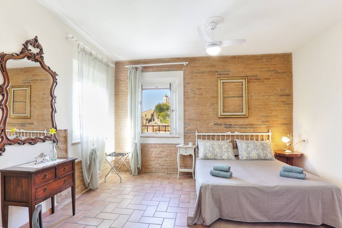 A bedroom with a brick wall, a large mirror, a bed with pillows, a bedside table with a lamp, and a window with curtains.