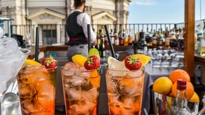 A bar with two glasses of iced drinks garnished with strawberries and lemon slices, a bartender in the background, and a view of a classical building.