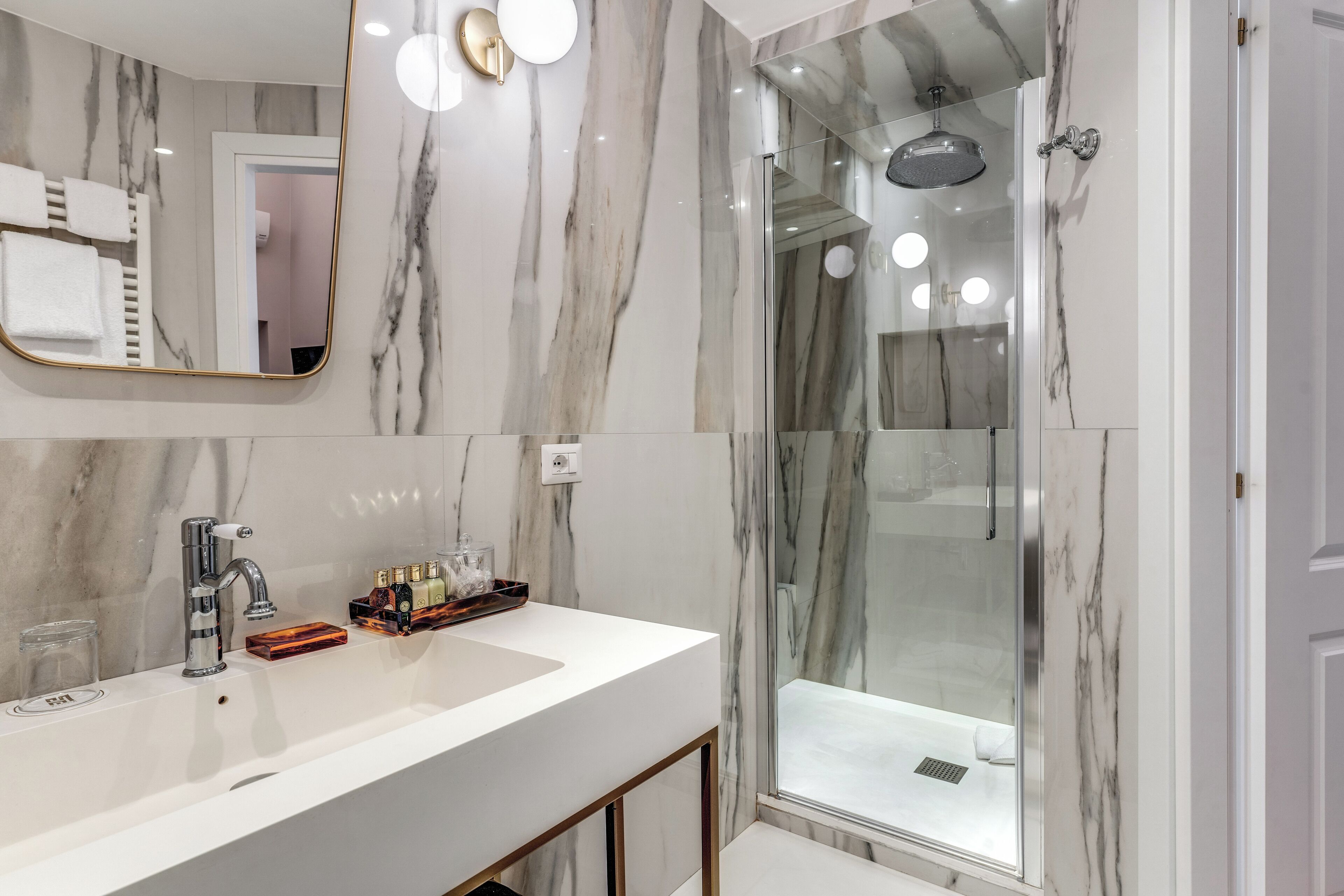 Un bany modern amb una paret de marbre, un mirall gran, un lavabo blanc i una dutxa envidriada.