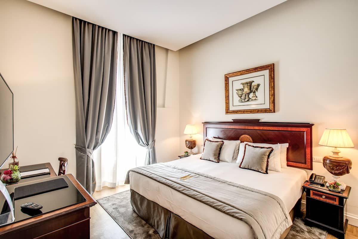 A hotel room with a large bed, bedside lamps, a wooden headboard, a desk with a computer, and a large window with curtains.