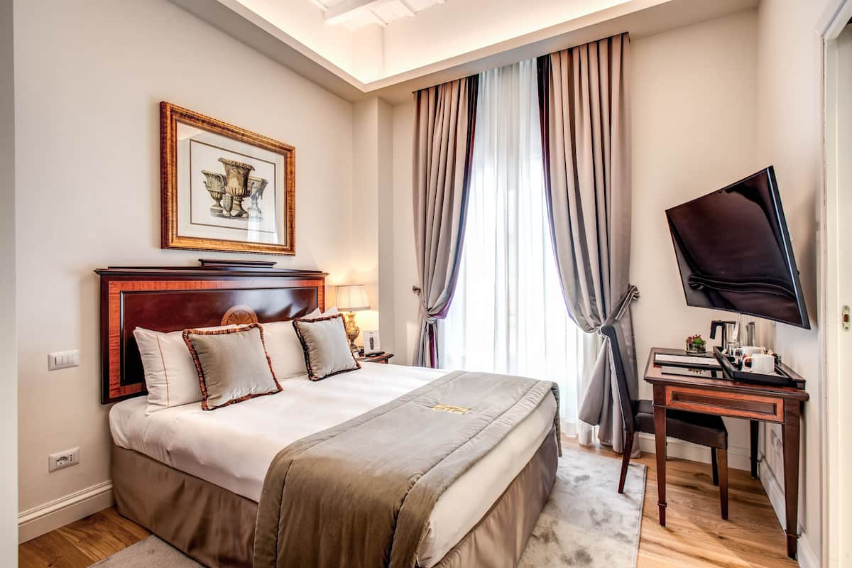A hotel room with a large bed, a desk with a TV, a framed picture on the wall, and a window with curtains.