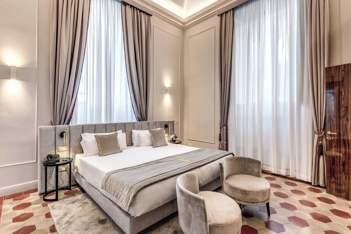 A modern hotel room with a large bed, a bedside table with a lamp, a chair, and a wooden door.