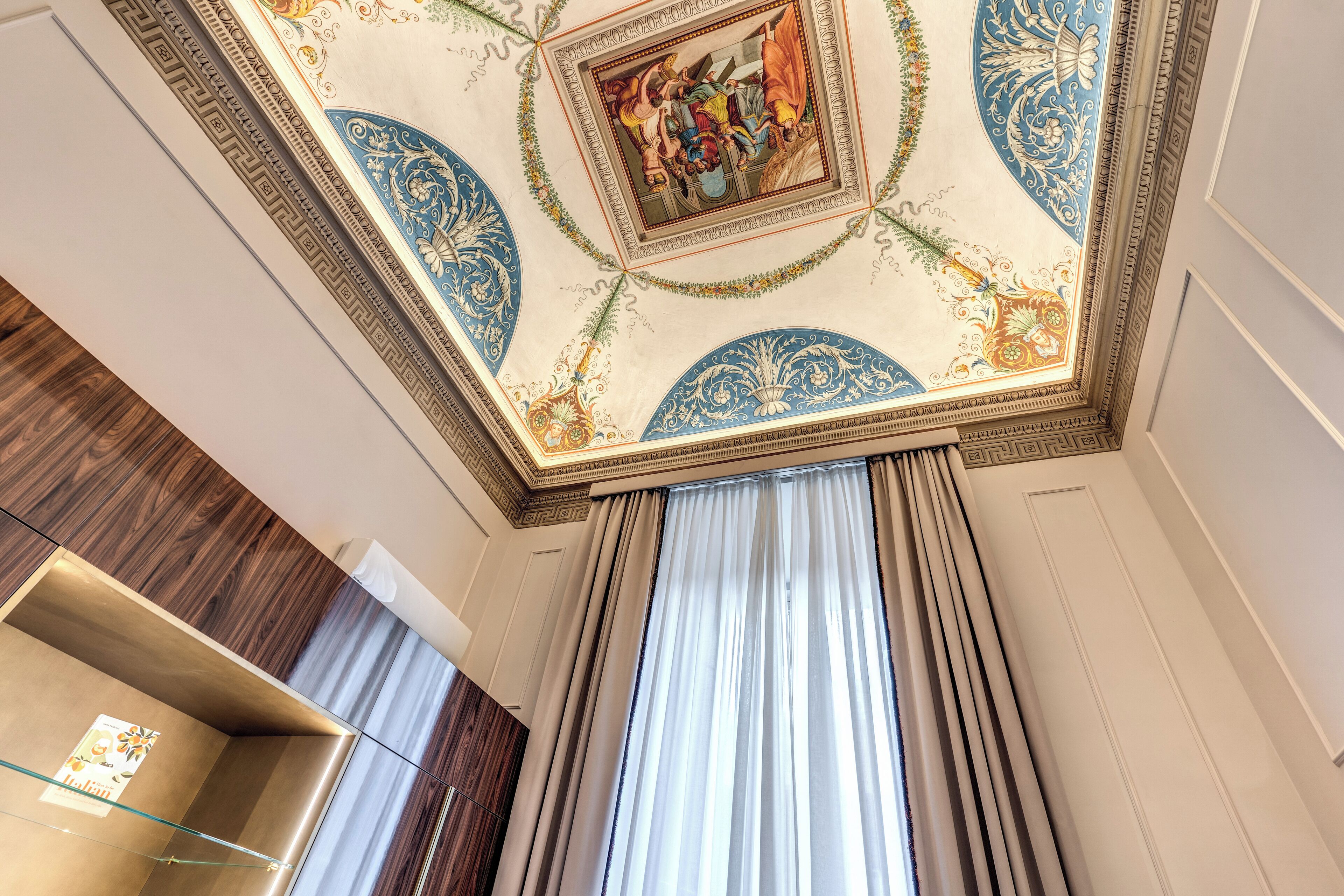 Une pièce dotée d’un plafond haut, orné de motifs décoratifs, de grandes fenêtres avec des rideaux et d’un meuble moderne en bois.