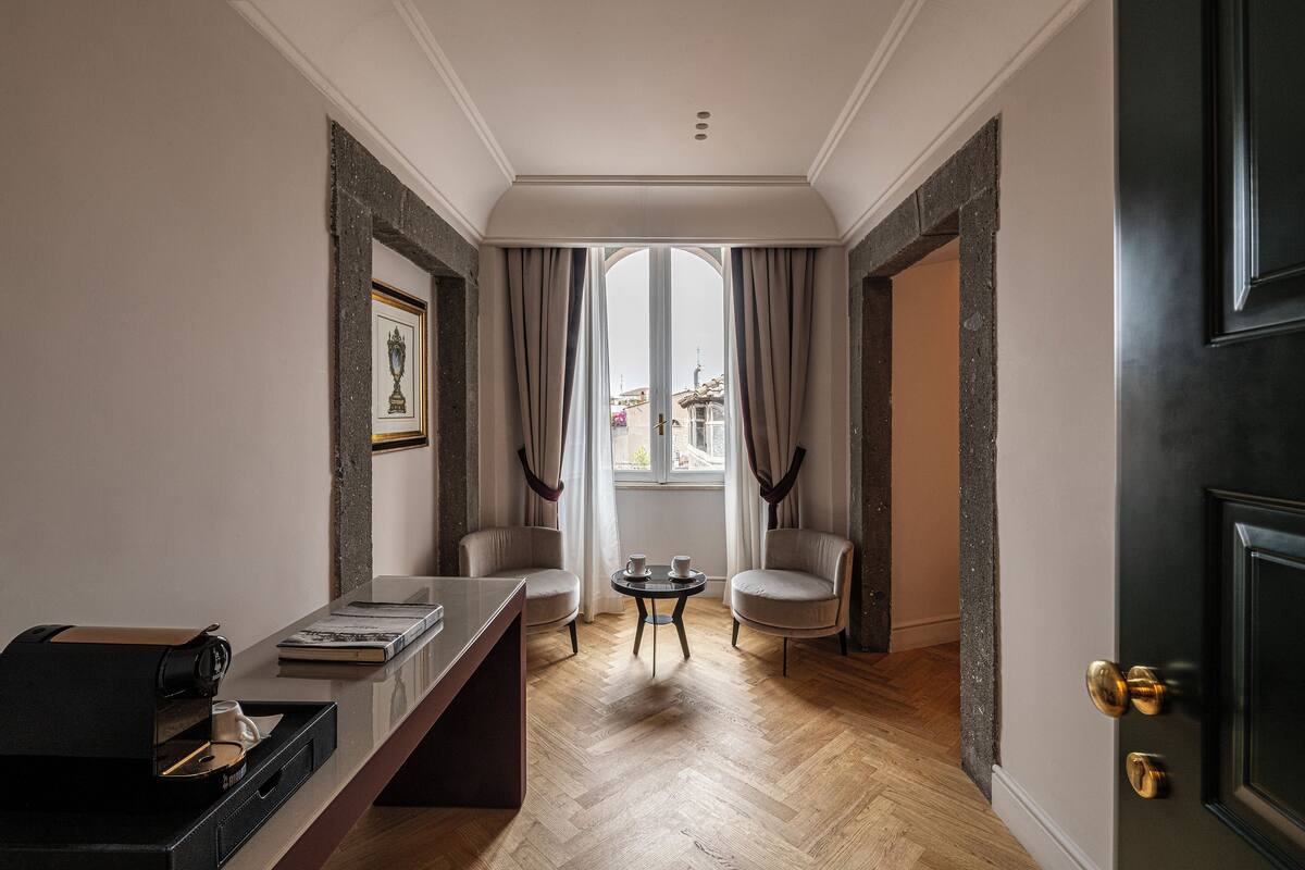 A room with a wooden floor, a desk with a printer, two chairs, a small table, and a large window with curtains.