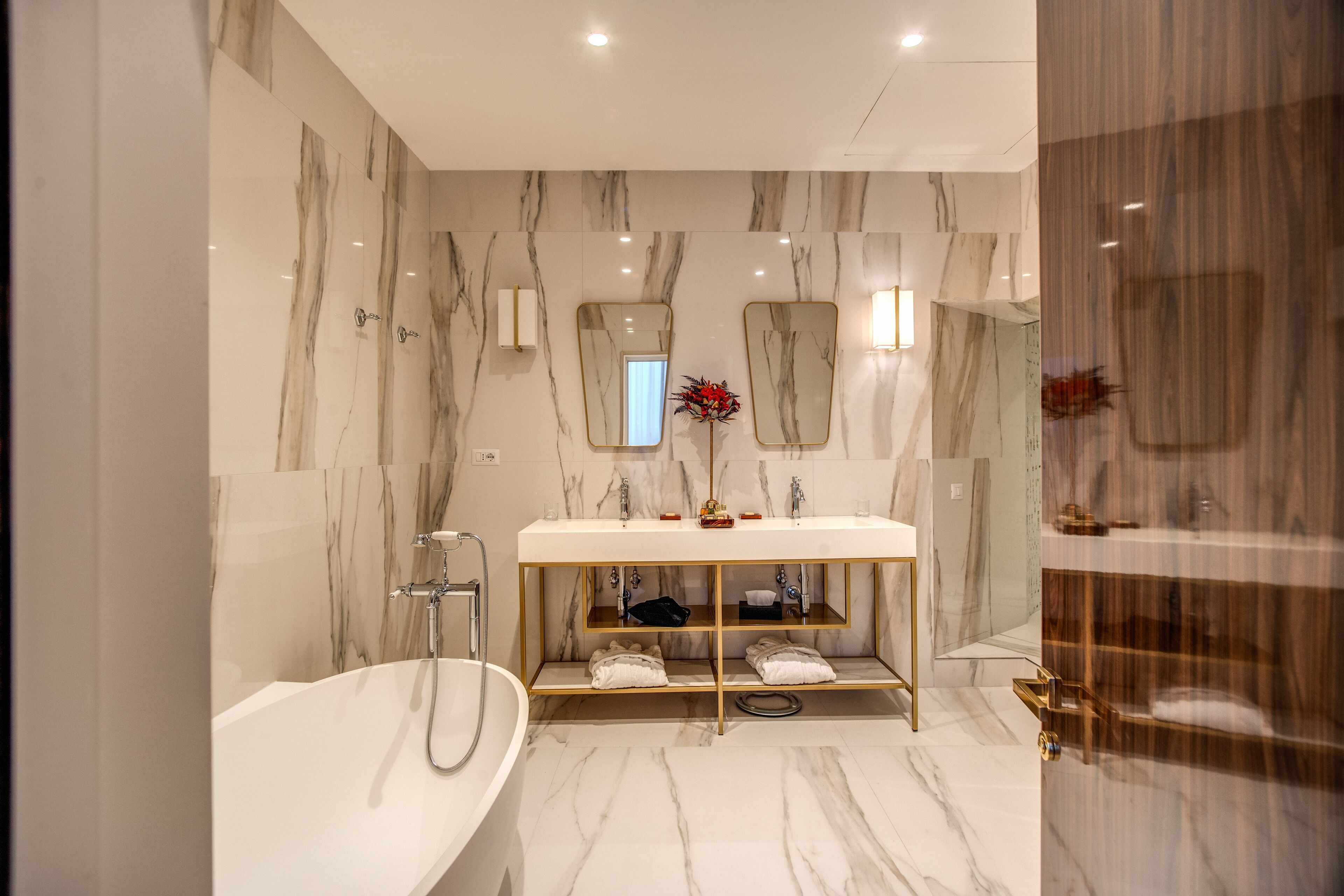 Ein modernes Badezimmer mit Marmorwand, einer freistehenden Badewanne, einem Waschtisch mit goldener Rahmung und einem Spiegel.
