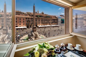 Μια θέα α πό το παράθυρο ενός δωματίου ξενοδοχείου, με θέα σε μια ιστορική πλατεία με ένα σιντριβάνι, κτίρια και ένα άγαλμα.