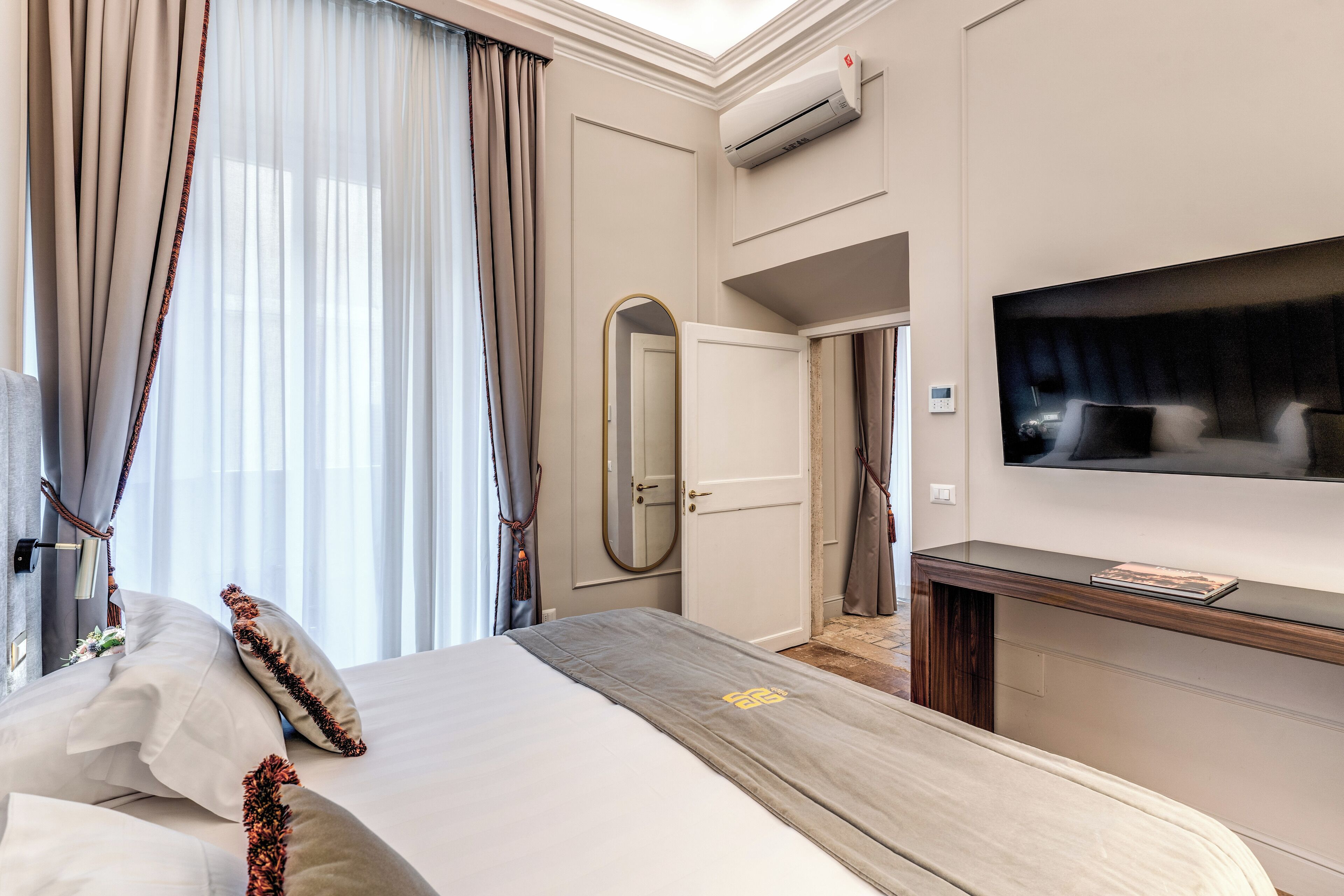 A hotel room with a bed, a TV, an air conditioning unit, and a door leading to another room.