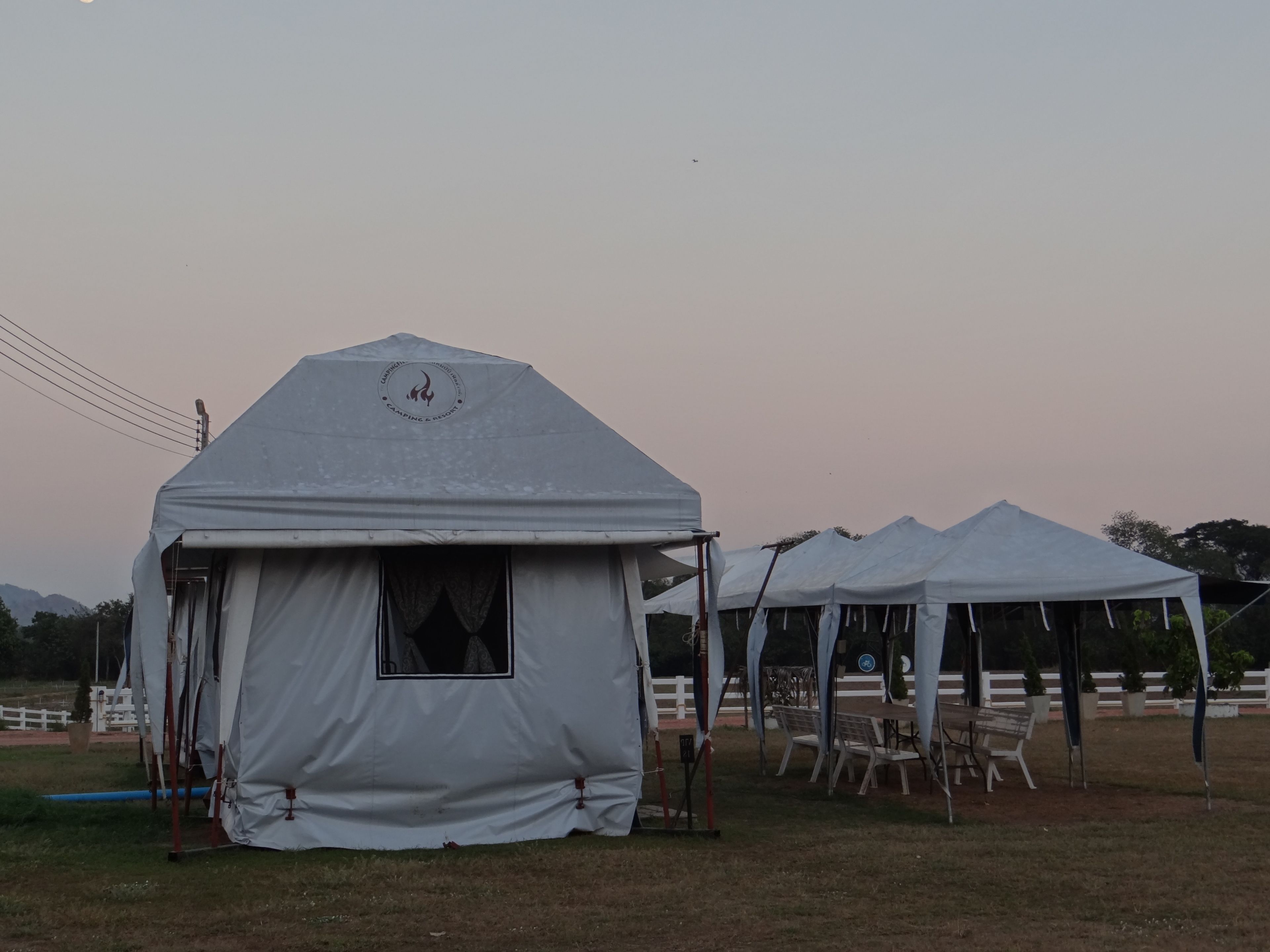 A series of white tents with a logo on the front, set up in an open field with chairs and tables arranged outside.