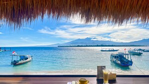 Une vue depuis une construction au toit de chaume, donnant sur une mer calme où flottent des bateaux, avec une montagne à l’horizon et une table en bois sur laquelle se trouvent une tasse et un verre.