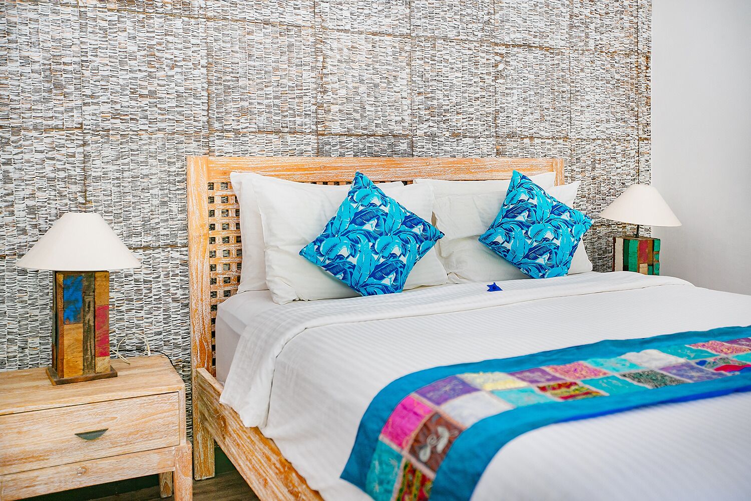 A bed with white bedding, two blue patterned pillows, a colorful quilt, and a wooden headboard. There are two bedside tables with lamps and a wall covered in a textured pattern.