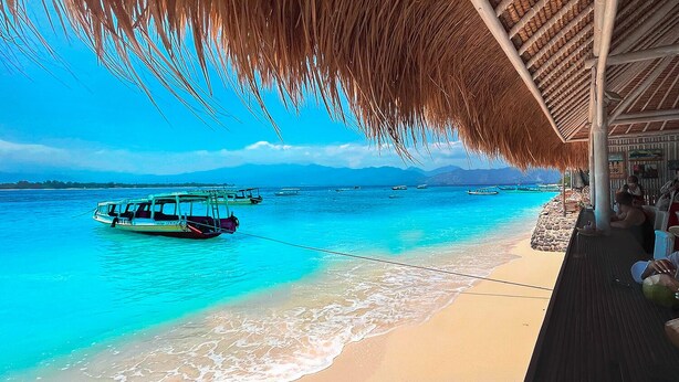 Une plage à l’eau turquoise et limpide, un bateau amarré près du rivage, et une structure à toit de chaume où sont assis des gens.