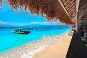 A beach with clear turquoise water, a boat docked near the shore, and a thatched-roof structure with people sitting. - Kura Kura Beach Resort (Gili Meno)