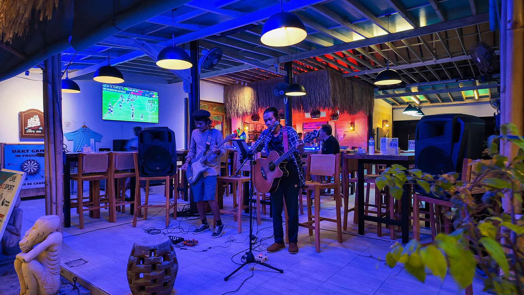 Un bar con arredi in legno e lampade a sospensione, uno schermo televisivo che mostra una partita sportiva e un musicista che si esibisce.