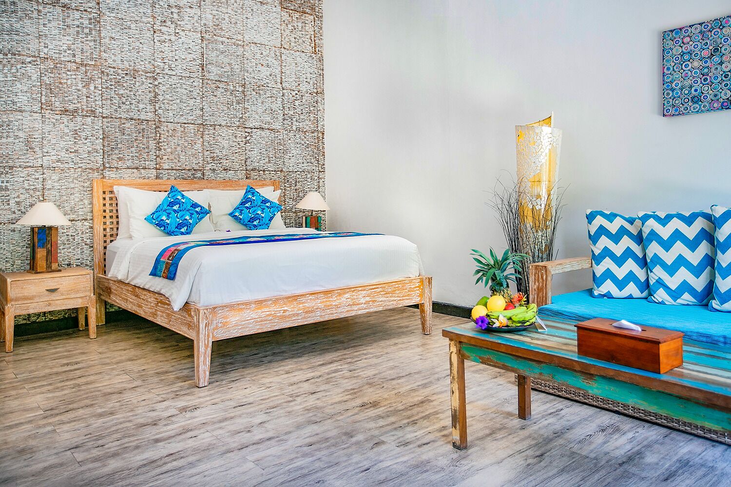 A bedroom with a wooden bed, two bedside tables, a nightstand, a bedside lamp, a small table with a bowl of fruit, and a colorful wall hanging.