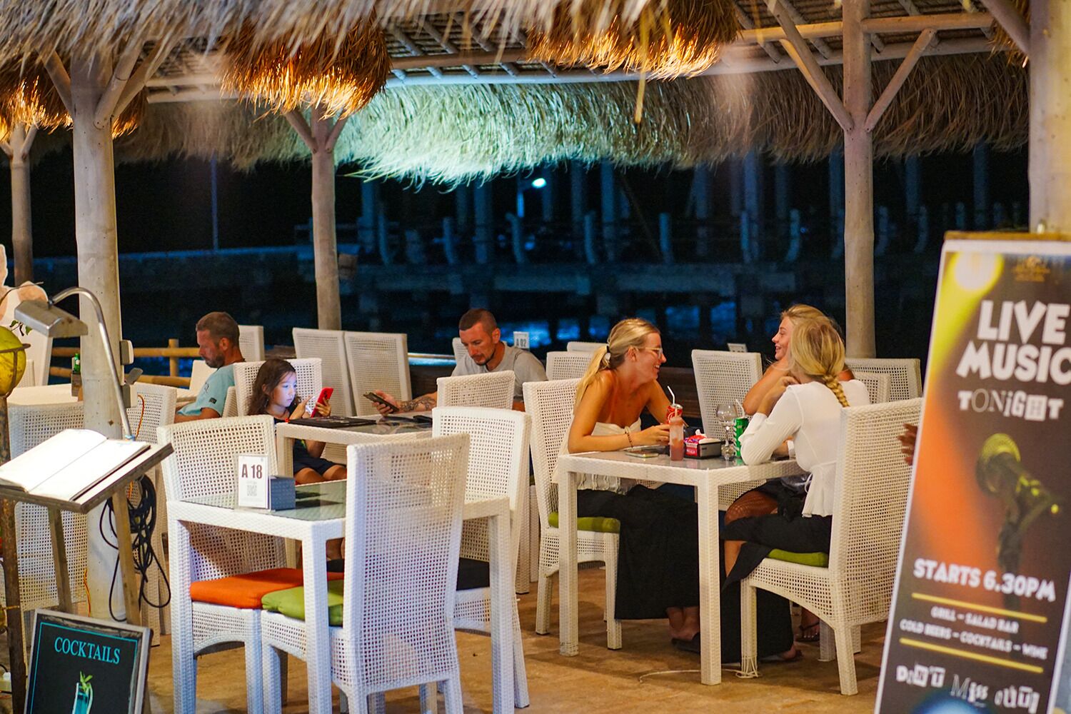 Un área de comedor al aire libre con sillas y mesas de mimbre, techo de paja y un cartel anunciando música en vivo a las 6:30 PM.
