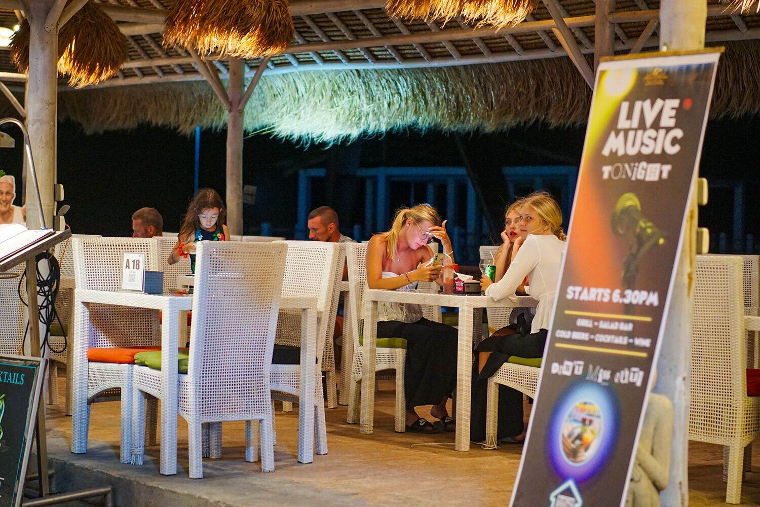 Un área de comedor al aire libre con sillas y mesas de mimbre, techo de paja y un cartel anunciando música en vivo a partir de las 6:30 PM.