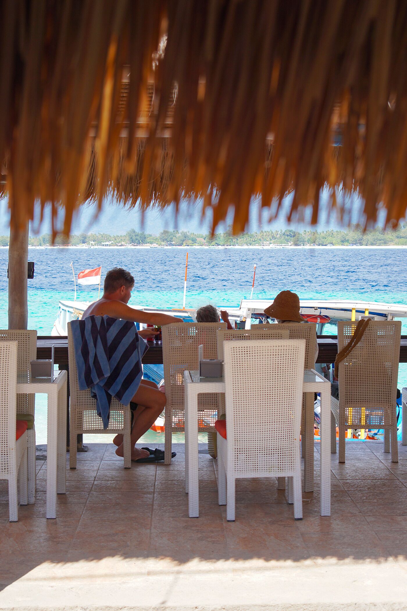 A covered outdoor seating area with white wicker chairs and a table, overlooking a body of water with boats.