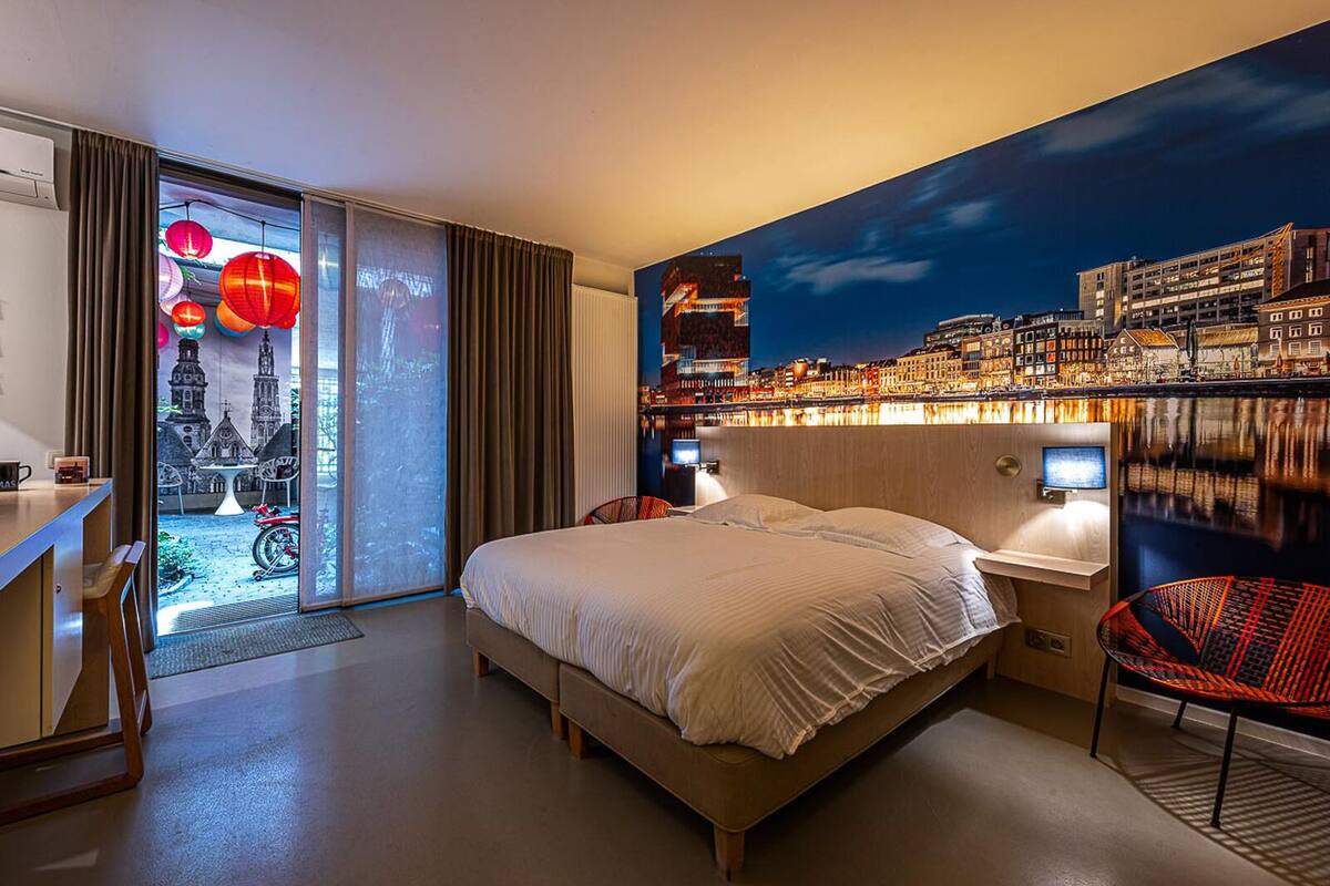 Habitación de hotel moderna con una cama grande, un escritorio y una silla. En la pared, un mural con un paisaje urbano y vistas de la ciudad a través de la ventana.