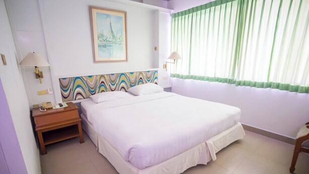 A hotel room with a large bed, a bedside table with a lamp, a wall with a colorful abstract painting, and a window with green curtains.