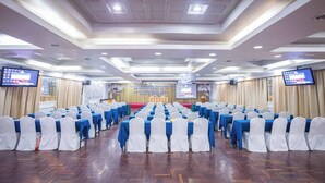 A conference room with rows of tables and chairs, a stage with a podium, and two screens on the walls.