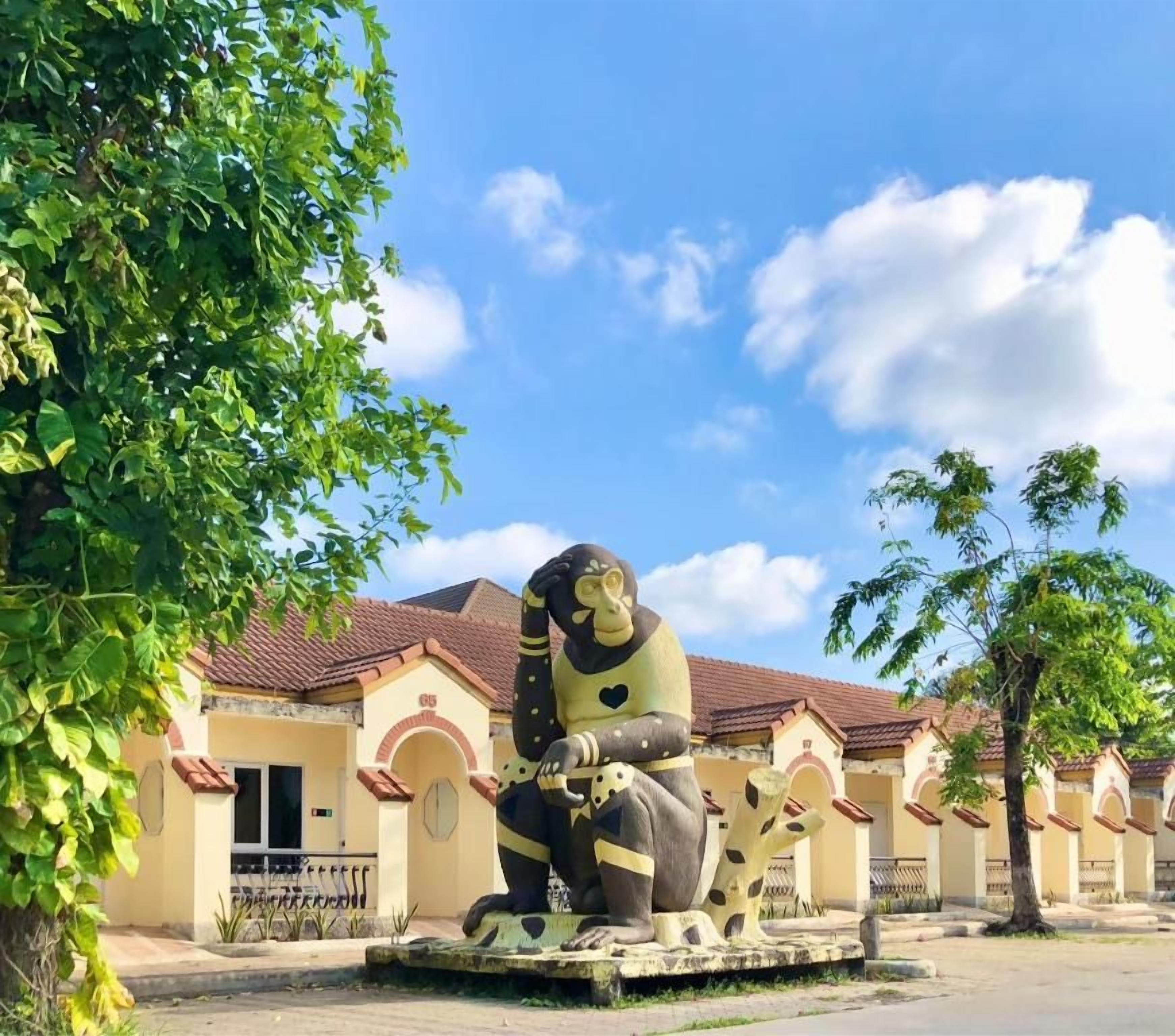 A large outdoor sculpture of a seated figure with a heart-shaped face, set against a building with a tiled roof and arched windows.
