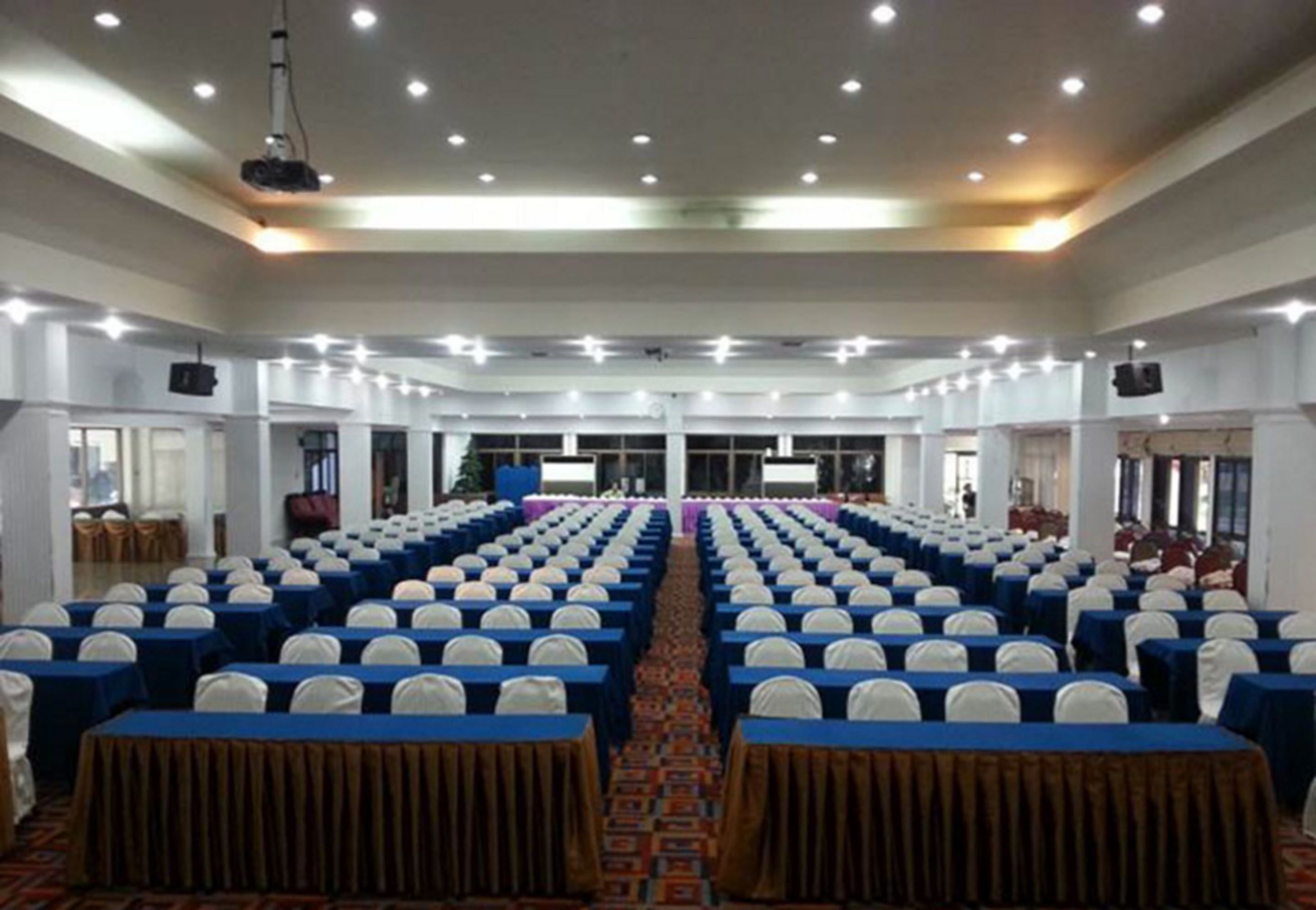 Un amplio salón de conferencias con filas de mesas y sillas, un escenario al frente y un proyector.