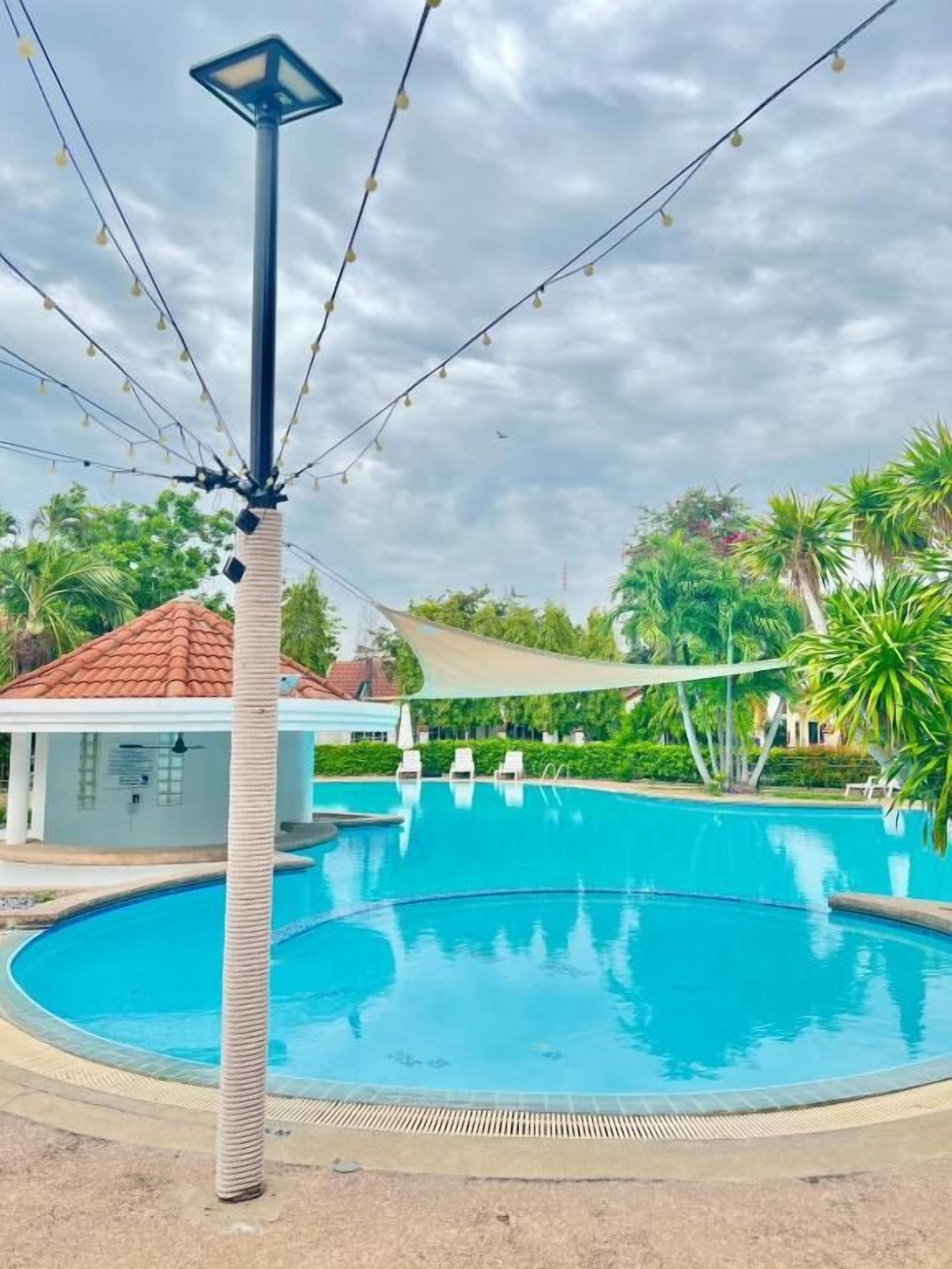 A swimming pool with a lamp post and string lights, surrounded by greenery and a building with a red roof.