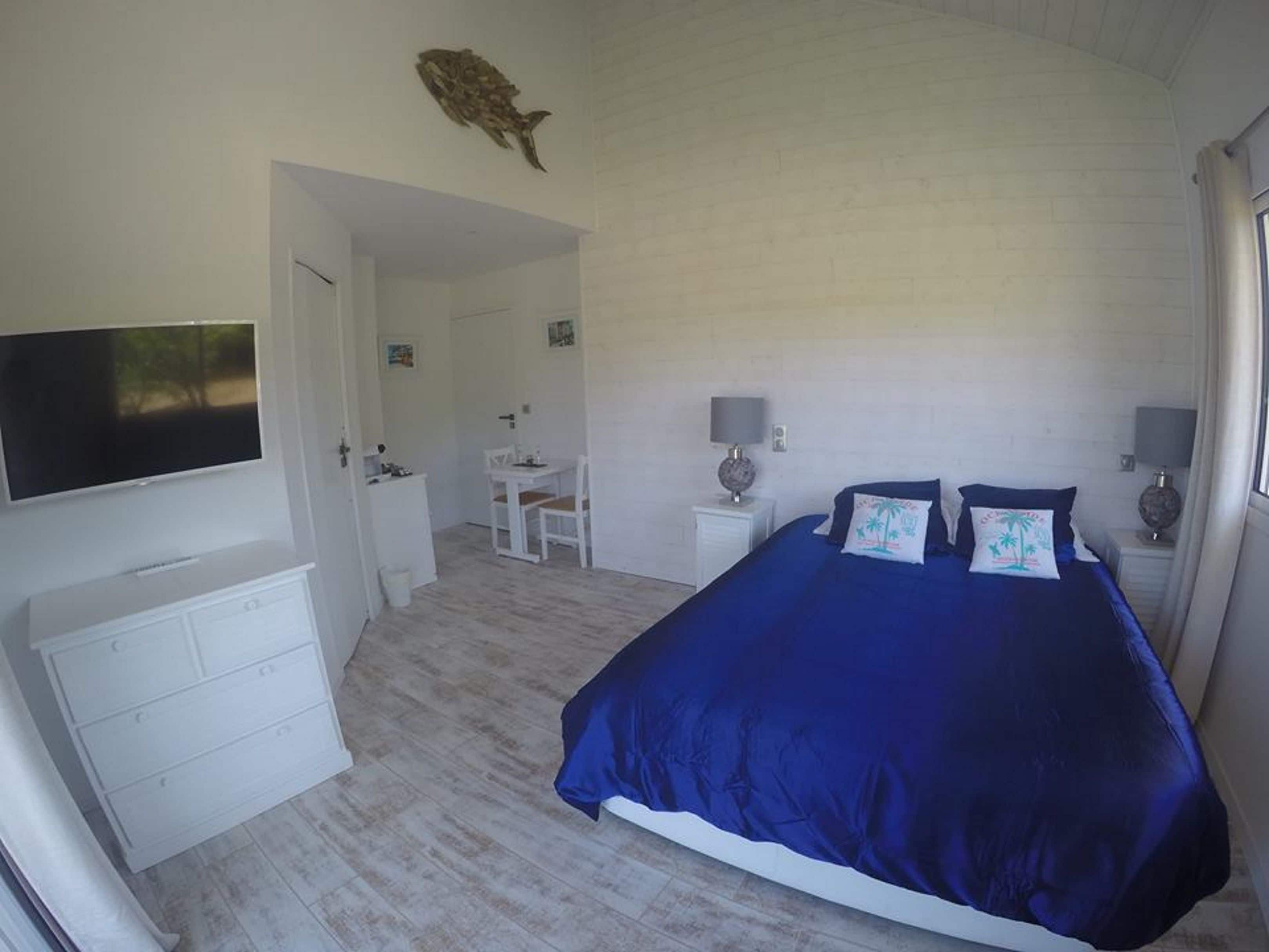 A bedroom with a large bed, a flat-screen TV, a desk with a chair, and a small table with a lamp.