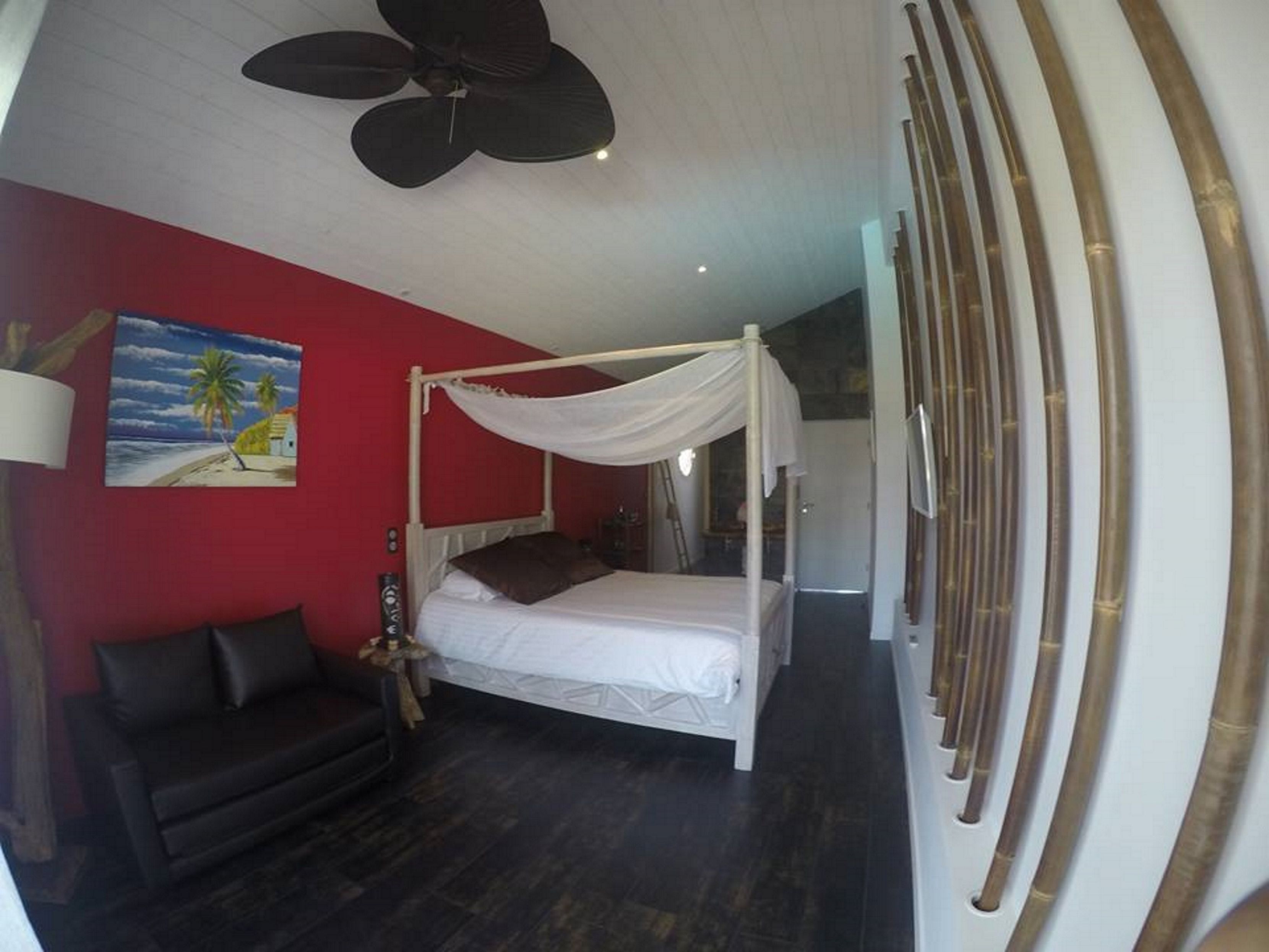 Un dormitorio con cama con dosel, un sofá de color oscuro, un ventilador de techo, un cuadro con escena de playa y un ventanal con persianas de bambú.