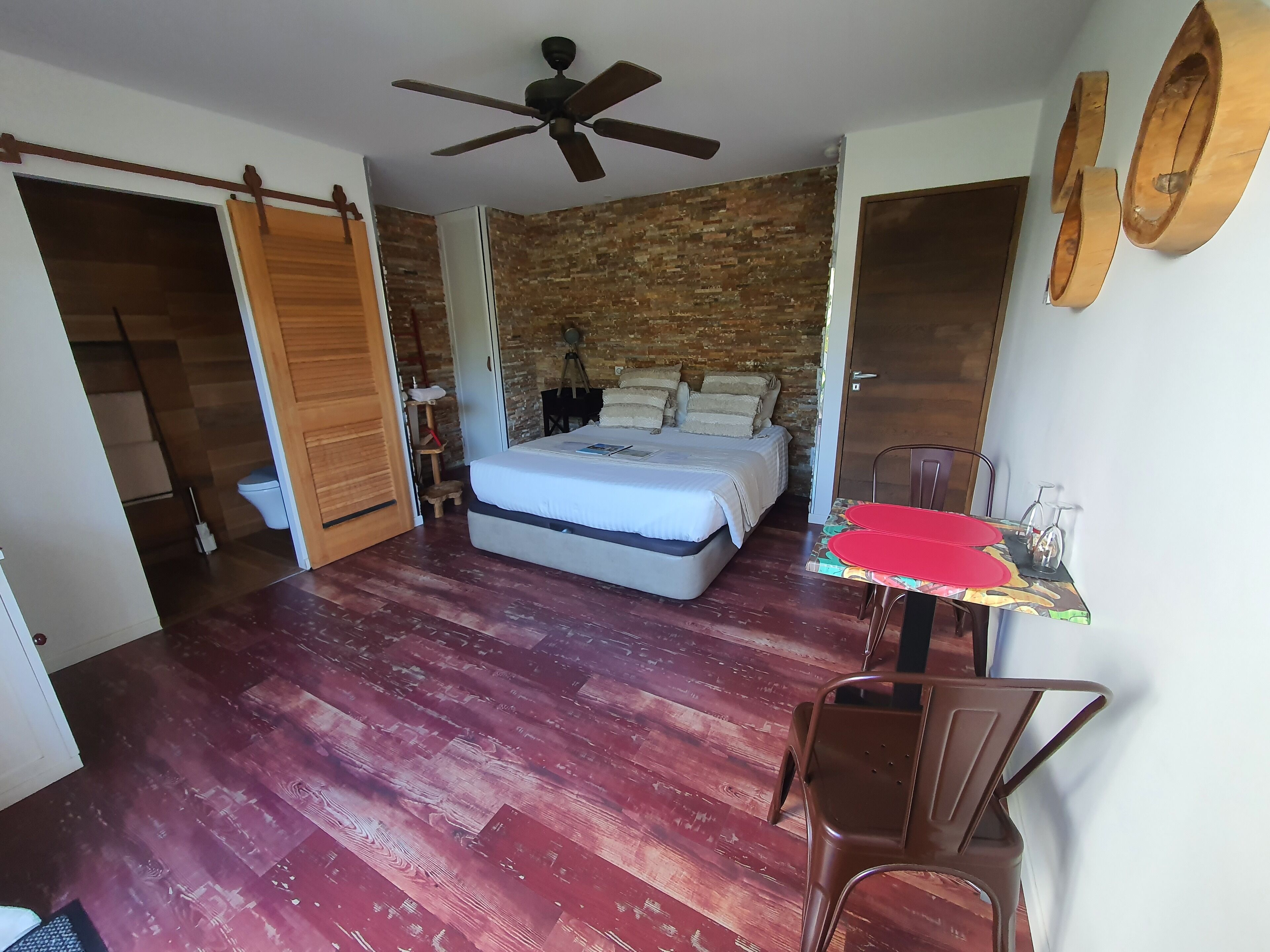A bedroom with a bed, a ceiling fan, a wooden door, and a table with chairs.