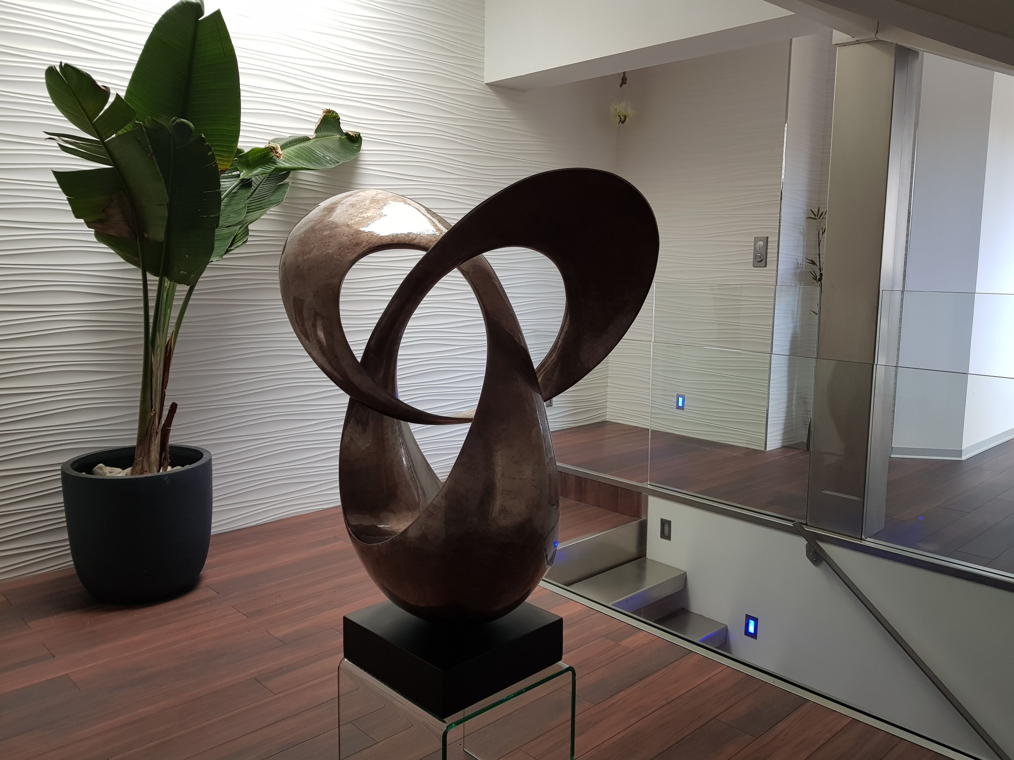 Una escultura moderna sobre un pedestal, una planta en maceta y una escalera con barandillas de vidrio.